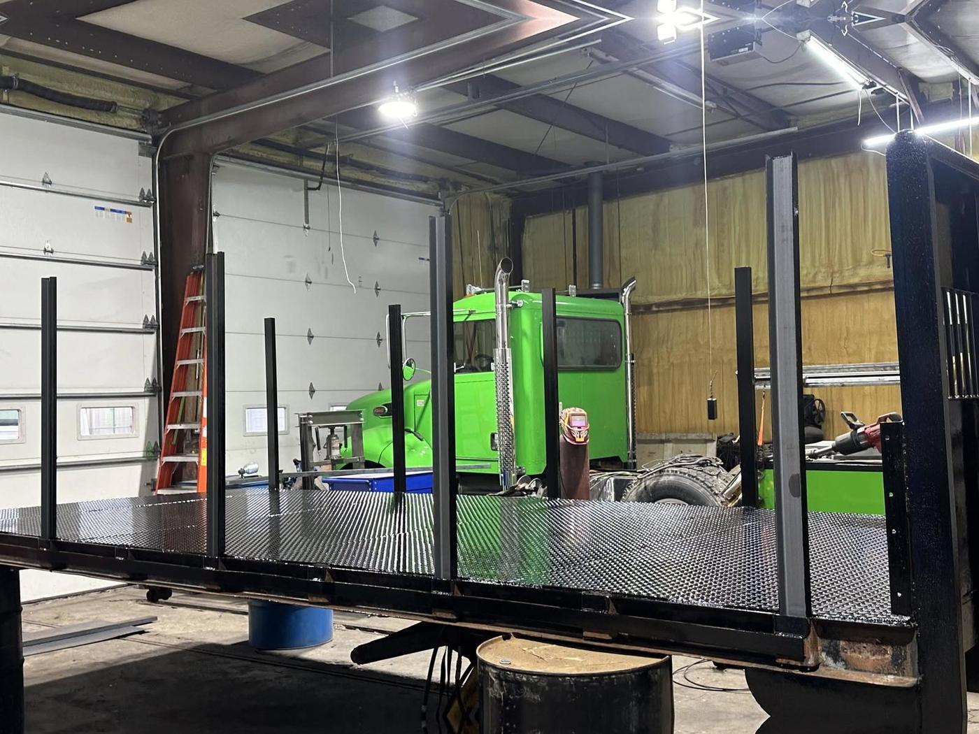 Freshly fabricated diamond-plate flatbed deck with stake posts, green semi tractor behind